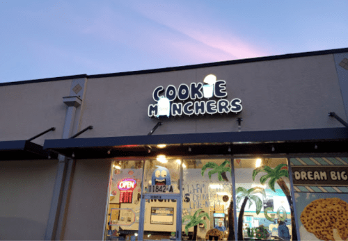 Sign for "Cookie Munchers" shop with a colorful storefront and palm tree decorations, illuminated at dusk.