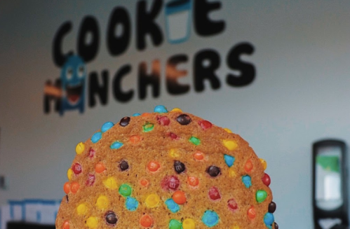 A colorful cookie with sprinkles held up in front of a "Cookie Crunchers" sign.