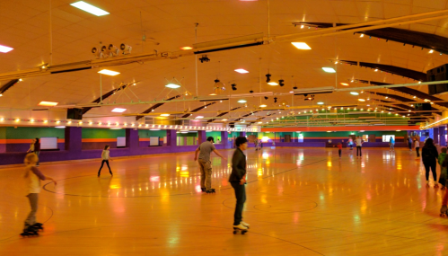 A brightly lit roller skating rink with people skating on a polished wooden floor. Colorful lights adorn the walls.