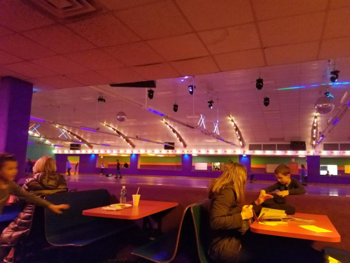 A colorful roller skating rink with people sitting at tables and others skating in the background under disco lights.