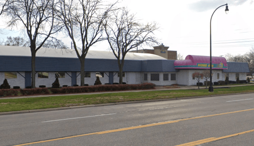 A large building with a colorful awning, surrounded by trees and shrubs, located along a street.