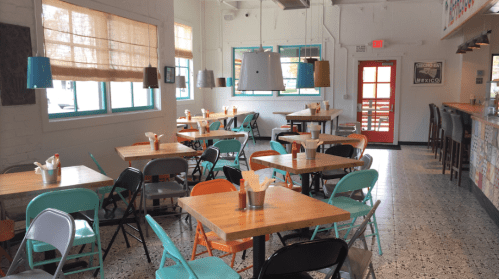 Bright, modern restaurant interior with wooden tables, colorful chairs, and a bar area, featuring large windows and pendant lights.