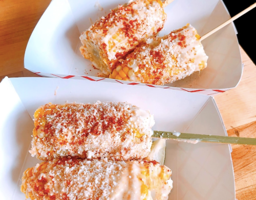 Two corn on the cob skewers topped with creamy sauce, cheese, and spices, served in paper trays.
