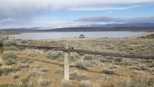 A scenic view of a lake surrounded by mountains, with a car parked near a wooden fence in the foreground.