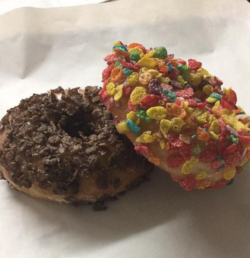 Two donuts on a white paper: one topped with chocolate sprinkles and the other with colorful cereal pieces.