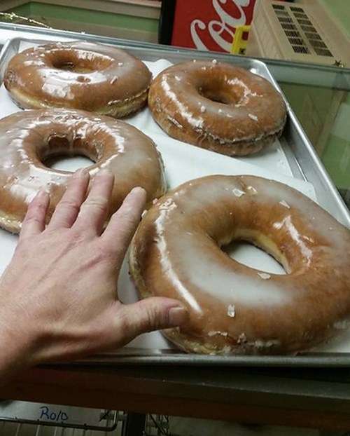 A hand reaches for a tray of large, glazed donuts, with four donuts displayed on a metal tray.
