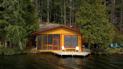 A bright yellow cabin on a lake, surrounded by trees, with a deck and chairs by the water.