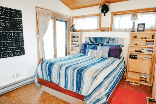 Cozy bedroom with a striped blue and white bedspread, wooden shelves, and large windows letting in natural light.