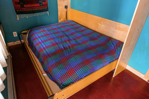 A wooden bed with a colorful checkered blanket in a room with teal walls and a patterned wall hanging.