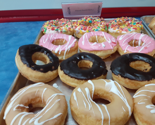 A tray of assorted donuts, including chocolate, pink frosted, and sprinkled varieties, with a sign in the background.