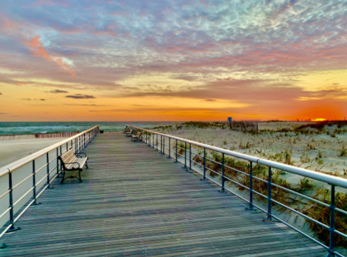 A wooden boardwalk leads to a beach at sunset, with benches and colorful clouds in the sky.