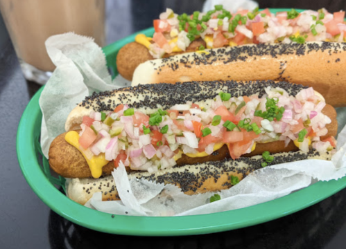 Two poppy seed hot dogs topped with diced tomatoes, onions, and green onions, served on a green plate.