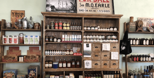 A wooden shelf displays various jars, bottles, and canned goods, with a vintage sign above advertising a sale.