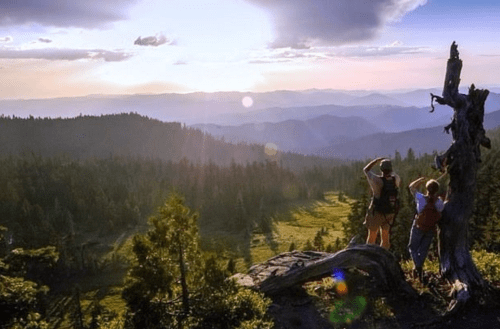 Two hikers stand on a rocky outcrop, gazing at a scenic mountain landscape during sunset.