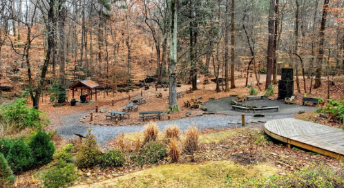 A serene forest clearing with picnic tables, a fire pit, and a small shelter surrounded by autumn foliage.