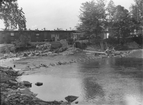 Black and white photo of a riverbank with rocks, trees, and buildings in the background; people are near the water.