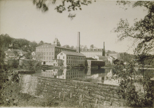 Historic industrial building by a river, featuring smokestacks and surrounding trees, in a sepia-toned photograph.