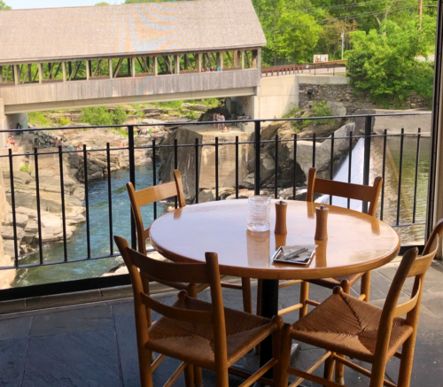 A table with chairs by a window overlooking a river and a covered bridge in a scenic outdoor setting.