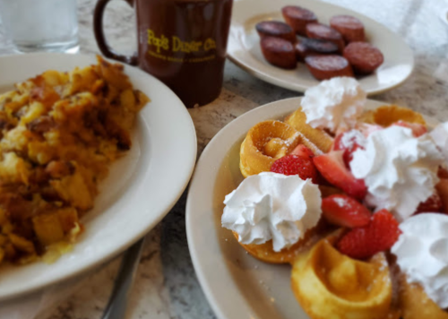 A plate of waffles topped with strawberries and whipped cream, alongside a serving of hash browns and a cup of coffee.