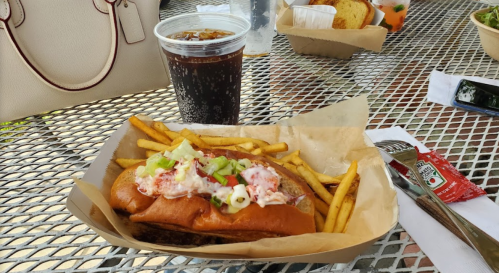 A lobster roll topped with vegetables, served with fries and a cup of soda on a table.