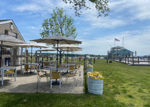 Outdoor dining area with tables and umbrellas, green grass, and a view of a waterfront building and American flag.