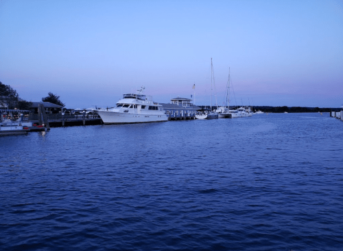 A calm marina at dusk with several boats docked and a soft blue sky reflecting on the water.