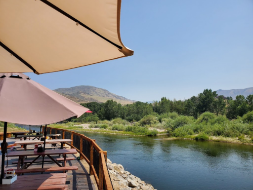 A scenic riverside view with wooden tables and umbrellas, surrounded by greenery and mountains under a clear blue sky.