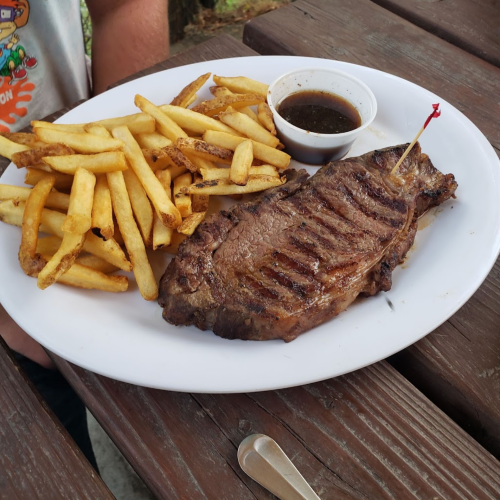 A plate with a grilled steak, fries, and a small cup of sauce, set on a wooden table.