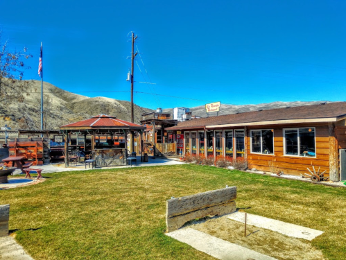 A rustic building with large windows, surrounded by grass and mountains, under a clear blue sky.