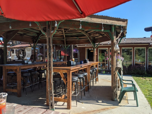 Outdoor bar area with wooden tables and stools, shaded by a red umbrella, surrounded by greenery and buildings.