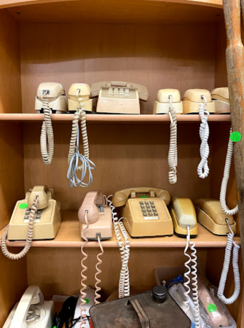 A wooden shelf displaying various vintage beige rotary and push-button telephones with coiled cords.