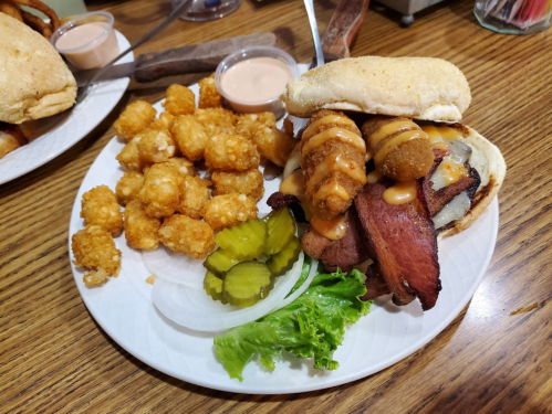 A plate with a sandwich featuring meat, pickles, lettuce, and tater tots drizzled with sauce on the side.