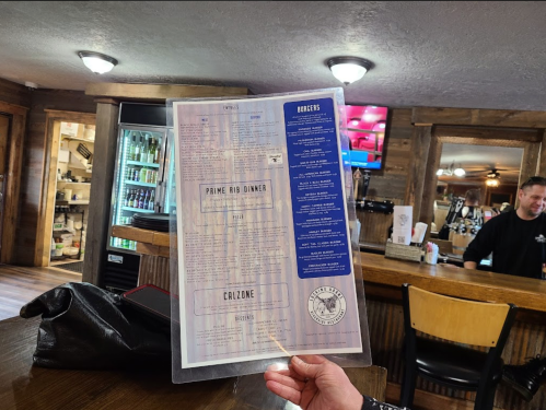 A person holds a menu featuring "Prime Rib Dinner" and "Calzone" in a rustic restaurant setting.