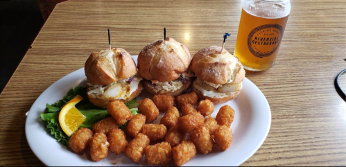 Three small sandwiches on a plate with tater tots and a glass of beer, garnished with lettuce and an orange slice.