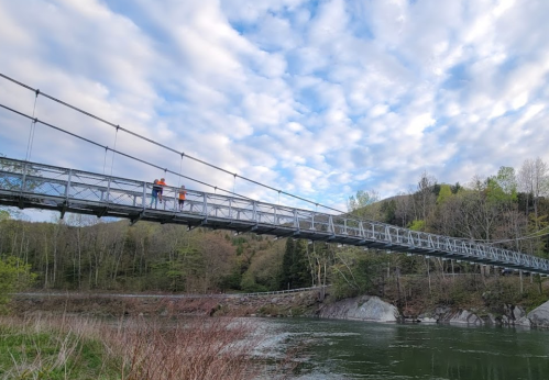 Two people walk across a suspension bridge over a calm river, surrounded by trees and a cloudy sky.