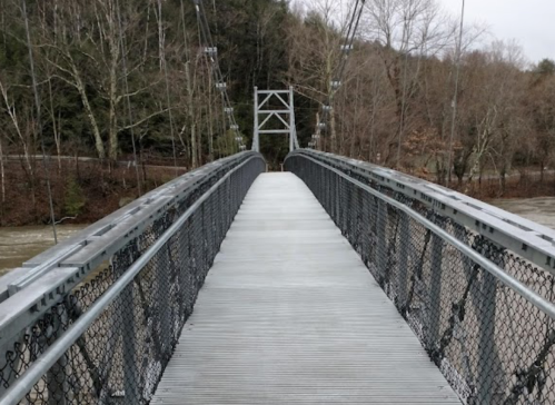 A suspension bridge with a wooden walkway, surrounded by bare trees and a river in the background.