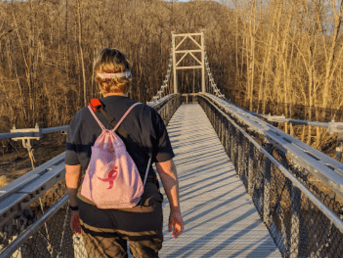 A person walks across a suspension bridge surrounded by bare trees, carrying a pink backpack.