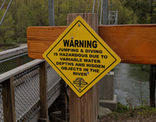Yellow warning sign about hazards of jumping and diving due to variable water depths and hidden objects in the river.
