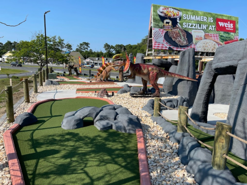 A mini-golf course featuring dinosaur sculptures, rocky terrain, and a summer promotional banner in the background.