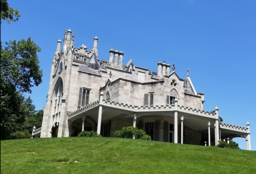 A grand stone mansion with intricate architecture, set on a green hillside under a clear blue sky.