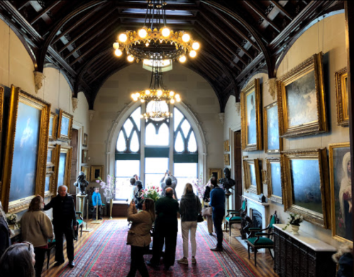 A grand hall with high ceilings, ornate chandeliers, and framed paintings, filled with visitors exploring the space.