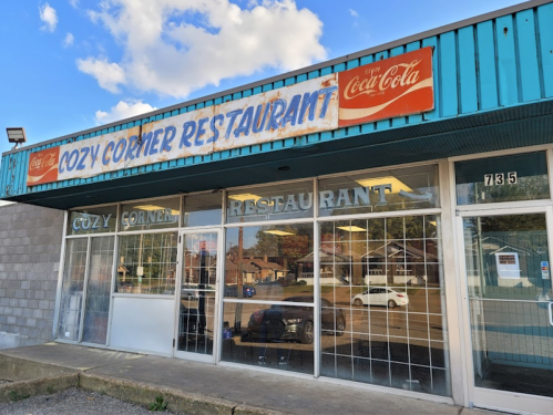 Exterior of Cozy Corner Restaurant with a vintage Coca-Cola sign, featuring large windows and a blue awning.