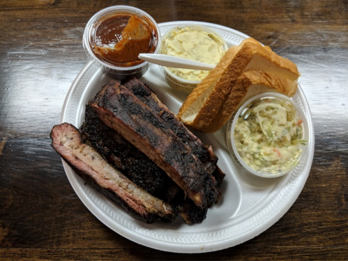 A plate with smoked ribs, barbecue sauce, two sides of coleslaw and potato salad, and slices of bread.