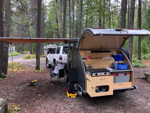 A compact camper trailer with an outdoor kitchen setup, parked in a wooded campsite with a vehicle nearby.