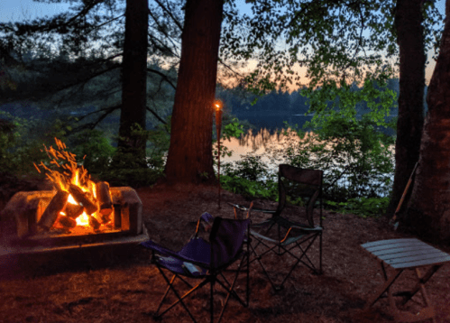 A cozy campfire surrounded by chairs near a tranquil lake at dusk, with trees framing the scene.