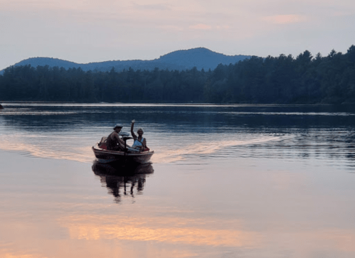 Two people in a small boat on a calm lake at sunset, surrounded by trees and mountains in the background.