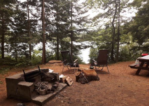 A serene campsite by a lake, featuring two chairs, a fire pit, and a picnic table surrounded by trees.
