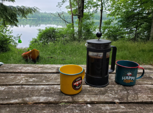 A French press and two colorful mugs sit on a wooden table, overlooking a serene lake surrounded by trees.
