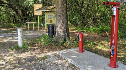 A bike repair station with tools, next to a trailhead sign and information kiosk in a wooded area.