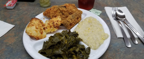 A plate of fried chicken, collard greens, grits, and cornbread, with utensils and a drink in the background.